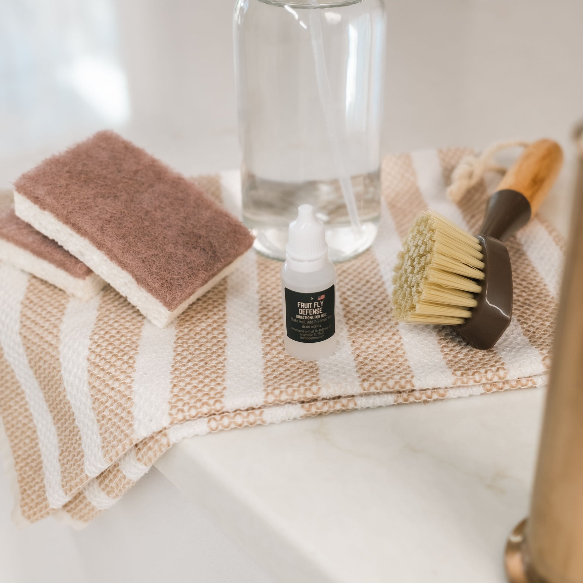 Cleaning supplies including a spray bottle, sponges, and a towel on a countertop. And Fruit Fly Defense sink drops.